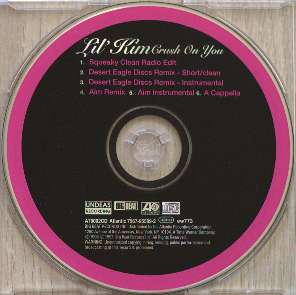 A CD disc from Lil' Kim's 'Crush On You' album, shown inside a clear plastic jewel case on a light wood surface. The disc has a black center with a bright pink ring and white and pink tracklist text.