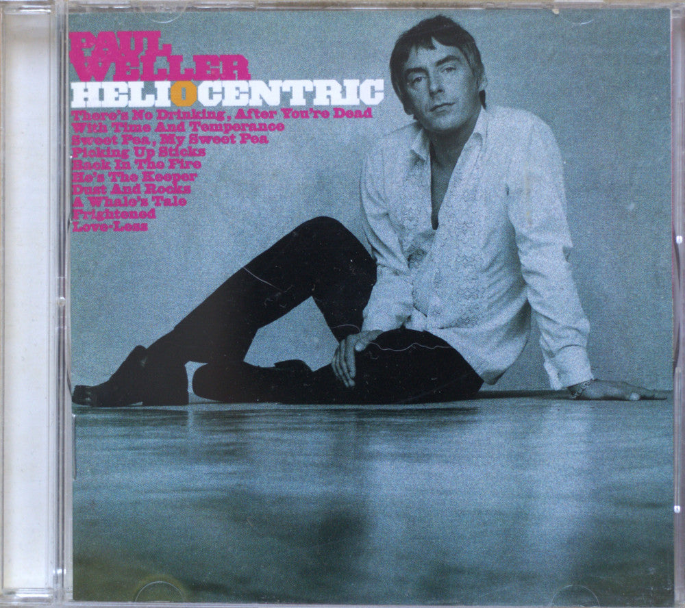 CD cover for Paul Weller' album 'Helio Centric', featuring a photo of the artist sitting on the floor wearing a white shirt and black trousers, with pink and white text on a blue-gray background.